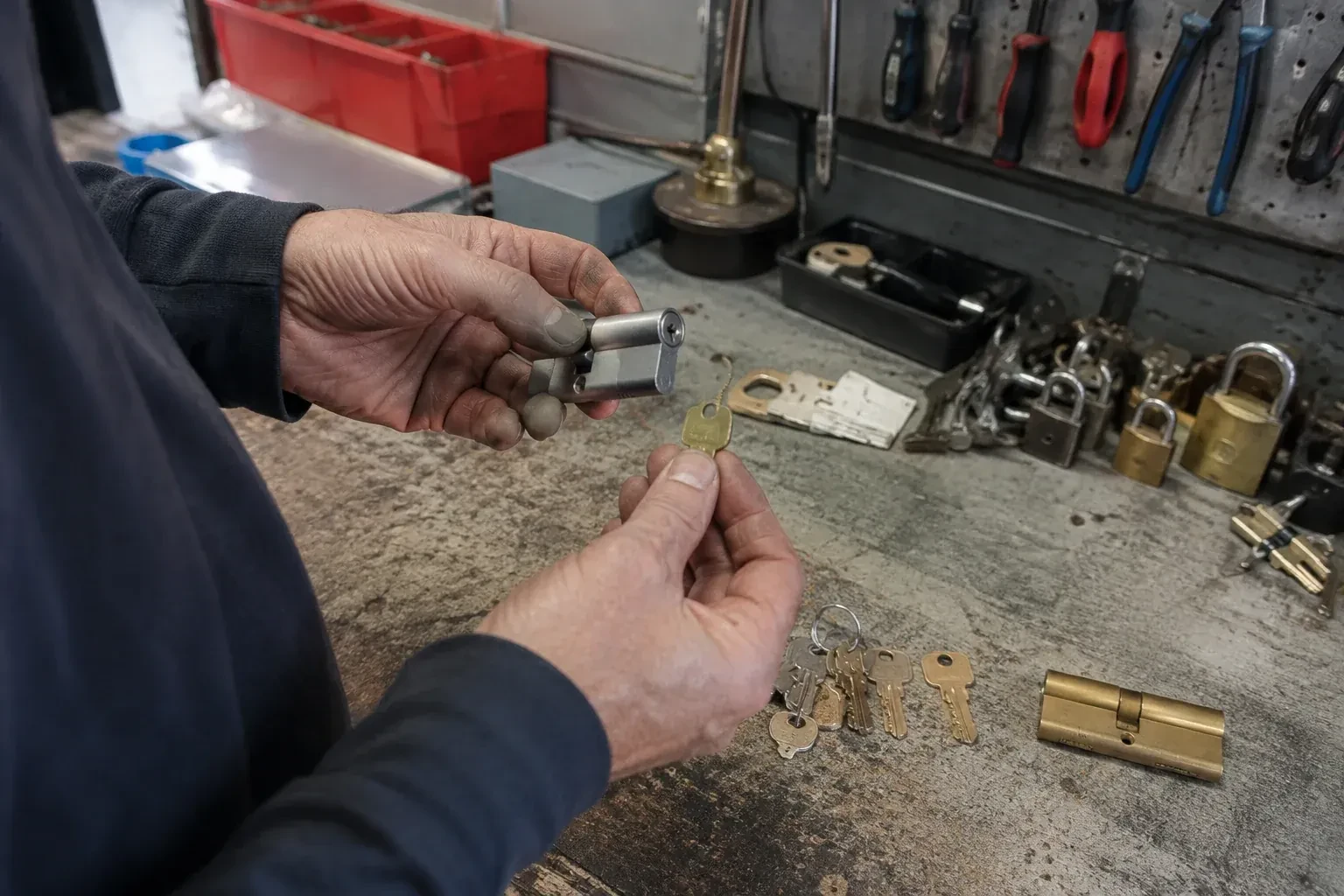 Hands holding a silver-colored lock cylinder and a brass-colored key above a workbench with padlocks, keys and tools. — Master Key System Design, Brampton